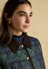 Geaca verde matlasată cu guler din catifea maro, nasturi aurii și un detaliu texturat al logo-ului pe piept. Puloverul cu guler înalt închis dedesubt.