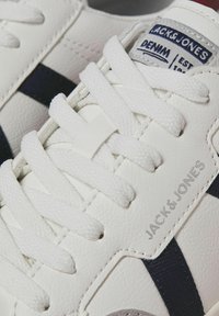 Witte leren sneaker met marineblauwe accenten, platte witte veters en een label met "JACK & JONES DENIM EST. 1990" in blauw en grijs.