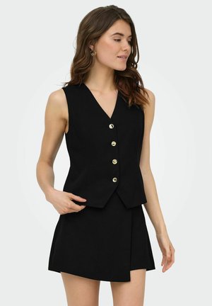 Femme portant un gilet noir sans manches avec des boutons dorés et une jupe noire assortie, debout et regardant sur le côté sur un fond uni.