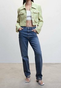 Zielona teksturowana krótka kurtka z dużymi guzikami, w połączeniu z niebieskimi jeansami o prostych nogawkach i białymi sandałami na paski.