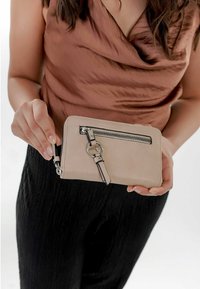 Portefeuille beige en matériau lisse, doté d'une poche zippée à l'avant avec des éléments en métal argenté et un détail de sangle. Tenue par une personne.