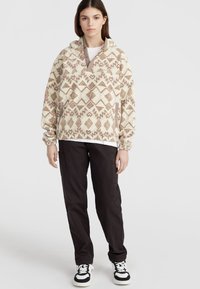 Fleece mikina s geometrickým vzorem v béžové a hnědé barvě, s polovičním zipem, velkou přední kapsou a elastickými manžetami, v kombinaci s tmavými kalhotami.
