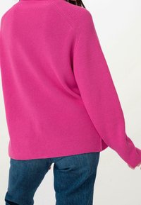 Pull en maille rose vif à finition texturée, présentant une coupe décontractée, des manches longues, et des fentes latérales, associé à un jean bleu foncé.