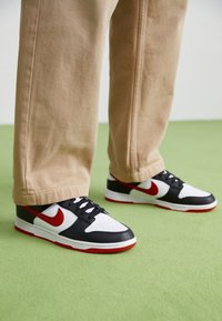 Nike Dunk-sneakers har en ovandel av svart läder, vit läder tån, röda detaljer och en röd sula. Kombinerade med khakibyxor på en grön yta.