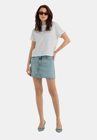 T-shirt bianco a maniche corte con grafiche stampate abbinato a una gonna di jeans blu chiaro. Gli accessori includono occhiali da sole marroni e scarpe con tacco blu.
