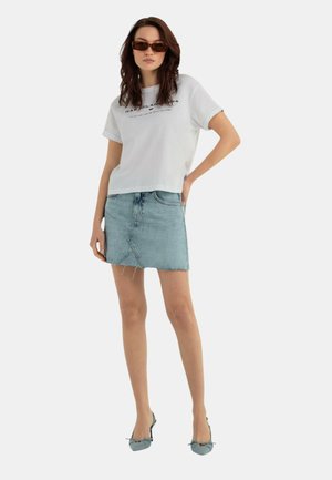 T-shirt bianco a maniche corte con grafiche stampate abbinato a una gonna di jeans blu chiaro. Gli accessori includono occhiali da sole marroni e scarpe con tacco blu.
