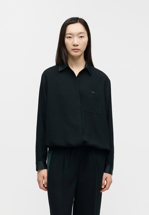 Femme debout devant un fond uni, portant une chemise noire ample à manches longues et un pantalon noir assorti, face à l'objectif avec une expression neutre.