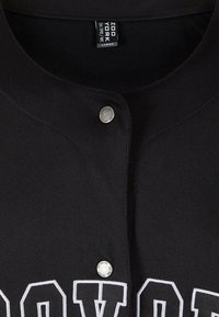 Maillot noir en tissu texturé, col rond et boutons-pression. Lettrage blanc sur le devant avec un détail accent brodé.