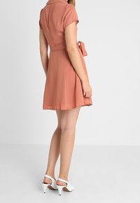 Vestito a maniche corte e colletto in rosa polveroso con una parte superiore attillata, gonna svasata e una cintura da annodare. Indossato con scarpe bianche con tacco alto.