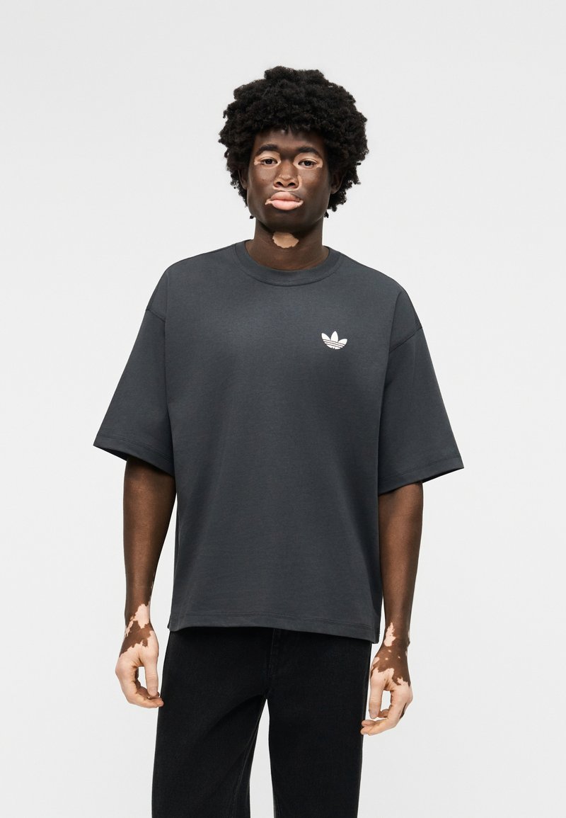 Junger Mann mit Vitiligo, der ein dunkelgraues, übergroßes Adidas-T-Shirt und schwarze Hosen trägt, vor einem schlichten hellen Hintergrund stehend.