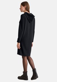 Svart huvtröja klänning med långa ärmar och detaljerad huva på baksidan. Bärs över svarta tights och kraftiga svarta stövlar, med en lös passform och knälång längd.