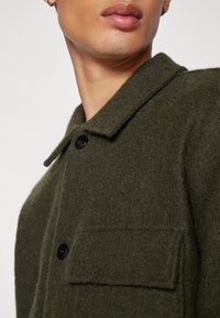 Olivgrön ullrock med texturfinish, som har en spetsig krage, svart knappstängning framtill och en bröstficka på vänster sida.