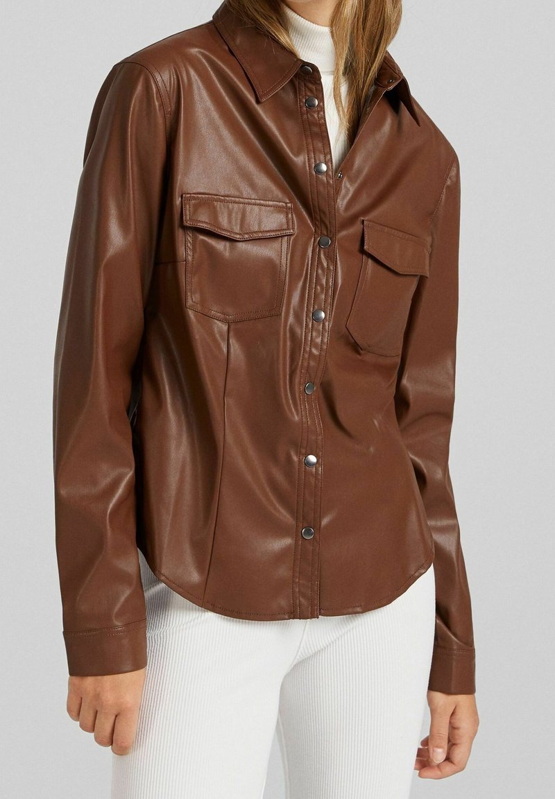 Brun konstläder skjorta med tryckknappar, en klassisk krage och två bröstfickor. Har en slät yta och en böjd nederkant.