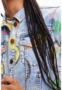 Jeansjacka i ljusblått tyg med färgglada grafiska tryck, bruna knappar och klassisk krage. Texturerad med synliga sömnadsdetaljer.