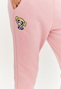 Ružové tepláky vyrobené z mäkkej látky majú na sebe nášivku s karikatúrou a text "VYROBENÉ Z CUKRU" v svetloružovej farbe pozdĺž strany.