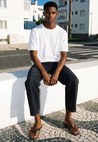 Jeune homme en T-shirt blanc, pantalon noir et tongs assis sur un bas mur blanc au bord de la rue avec des bâtiments modernes derrière.