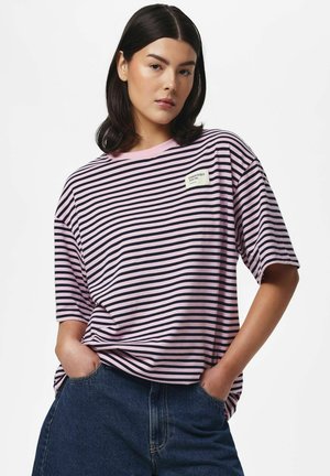 Ung kvinde med mørkt hår står med hænderne i lommerne iført en pink og sort stribet t-shirt og blå jeans mod en ensfarvet baggrund.