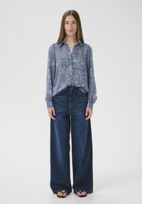Modrá košile s dlouhými rukávy a vzorem, nošená s riflovými kalhotami s širokými nohavicemi a vysokým pasem. Černé sandály doplňují celý outfit.