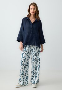 Chemise en lin marine avec col et boutons, associée à un pantalon blanc orné de motifs abstraits bleus, stylisée avec des baskets blanches.