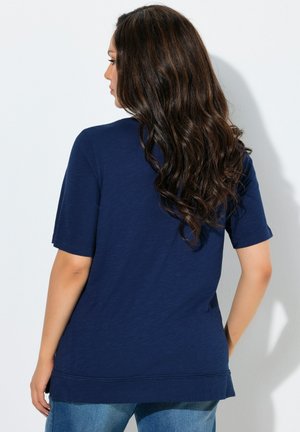 Kvinde med langt, bølget brunt hår iført en enkel navyblå kortærmet skjorte og blå jeans, vendt væk mod en hvid baggrund.