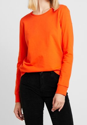 Mujer con una camiseta de manga larga de color naranja brillante y pantalones negros, de pie frente a un fondo gris claro liso.