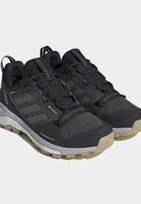 Svarte atletiske sko med teksturert mesh-overdel, tre grå striper, forsterket tå og beige såle. Har GORE-TEX-merke for vanntetting.
