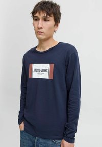 Fiatal férfi rövid barna hajjal, sötétkék hosszú ujjú pólót visel, rajta a "JACK & JONES" logó, kék farmert hord, egy sima háttér előtt áll.