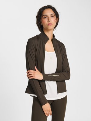 Kobieta o ciemnych kręconych włosach, ubrana w brązową kurtkę na suwak na białym topie, stojąca z założonymi ramionami na jednolitym białym tle.