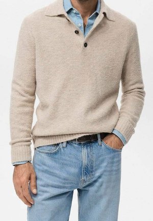 Uomo che indossa un maglione beige con bottoni sopra una camicia di jeans azzurro chiaro e jeans azzurri chiari, con una mano in tasca.