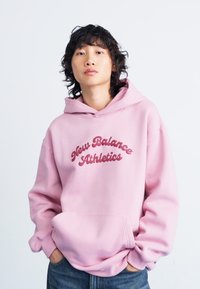 Haut à capuche rose avec poche avant et cordon de serrage, arborant le texte "New Balance Athletics" dans une teinte plus foncée de rose. Texture en tissu douce.
