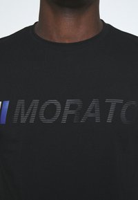Černé tričko vyrobené z bavlny má grafický design se slovem "MORATO" v různých odstínech šedé a modrým akcentem vlevo.