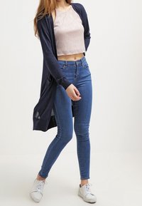 Námořnický dlouhý cardigan přes světle růžový crop top, v kombinaci s modrými skinny džíny a bílými teniskami. Měkká tkanina, přiléhavý střih a kapsy.