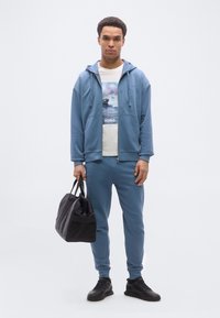 Νεαρός άνδρας με γαλάζιο φούτερ με κουκούλα και φόρμες κρατάει μαύρη τσάντα duffel, φοράει λευκό μπλουζάκι με σχέδιο και μαύρα αθλητικά παπούτσια, στέκεται μπροστά σε απλό φόντο.