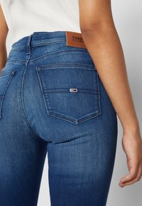 Jeans de mezclilla azul ajustados con una textura suave, cintura de altura media, bolsillos traseros dobles y una etiqueta de cuero marrón en la cinturilla.