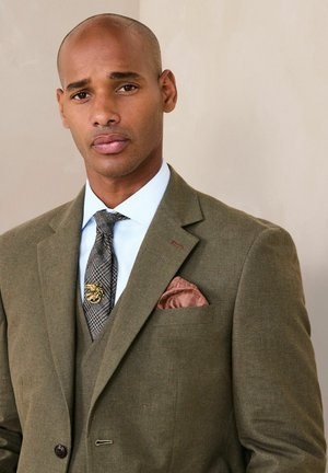 Mann mit rasiertem Kopf, der einen olivgrünen Anzug, ein blaues Hemd, eine gemusterte Krawatte mit goldener Nadel und ein braunes Einstecktuch trägt, vor neutralem Hintergrund.