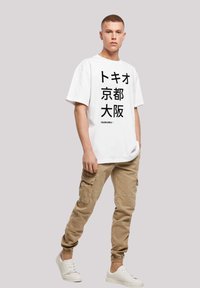 White graphic t-shirt with Japanese text, beige cargo pants with pockets, and white sneakers. Model stands against a neutral background.