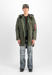 Νεαρός άνδρας που φοράει πολύκλωνο πράσινο στρατιωτικού στυλ μπουφάν, καπέλο παραλλαγής, ξεθωριασμένα τζιν και μαύρες μπότες, στέκεται μπροστά σε λευκό φόντο.