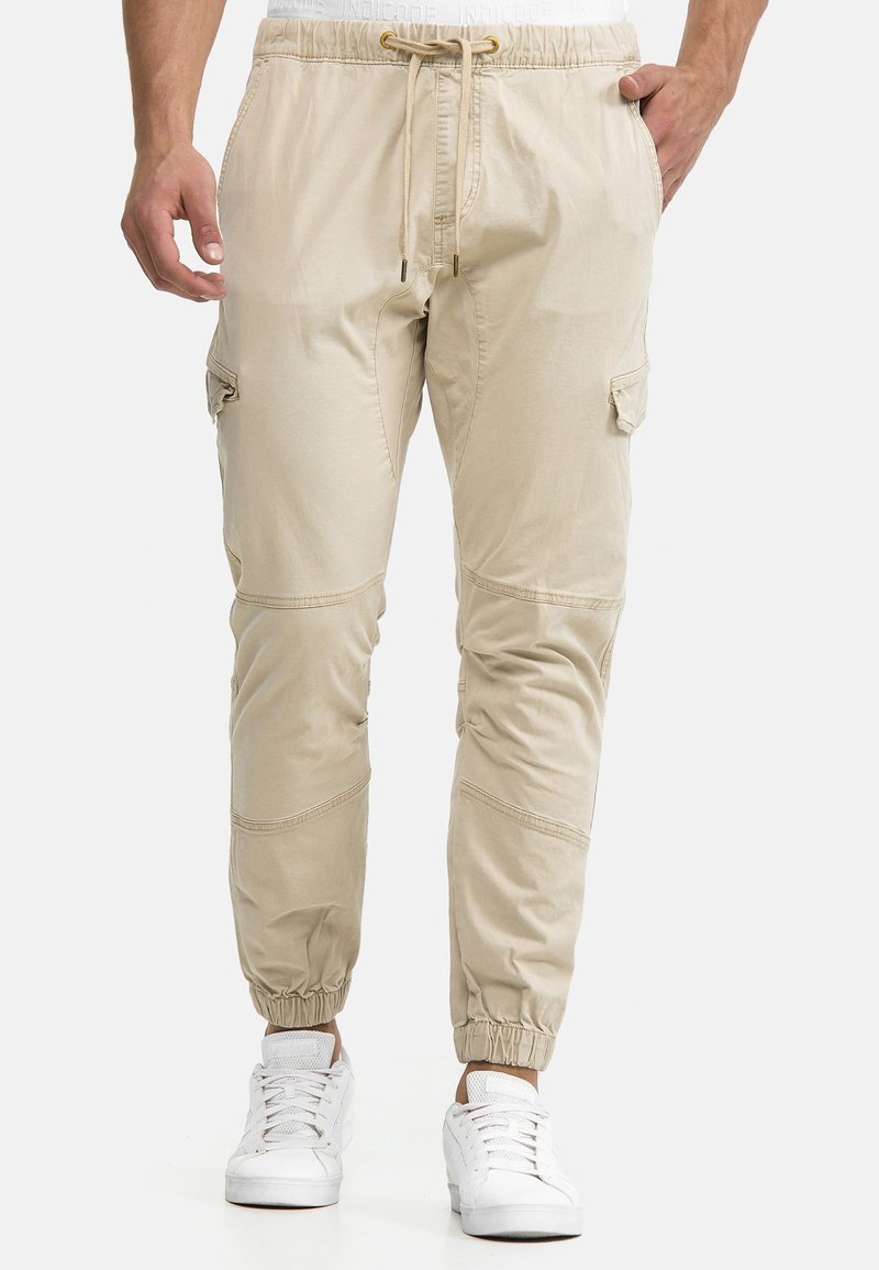 Homme portant un pantalon jogger cargo beige avec bas élastiqués, cordon à l'avant, poches latérales et baskets blanches, debout sur un fond uni.