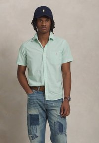 Camicia a maniche corte a righe verde chiaro con taschino sul petto a sinistra, indossata con jeans blu strappati e un berretto blu navy.