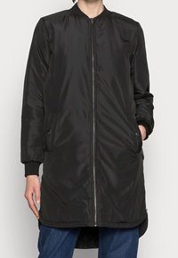 Personne portant une veste bomber noire longue à fermeture éclair avec des poches, des poignets côtelés et un jean bleu, debout devant un fond clair.