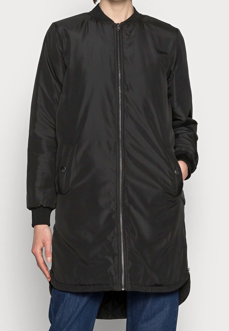 Personne portant une veste bomber noire longue à fermeture éclair avec des poches, des poignets côtelés et un jean bleu, debout devant un fond clair.