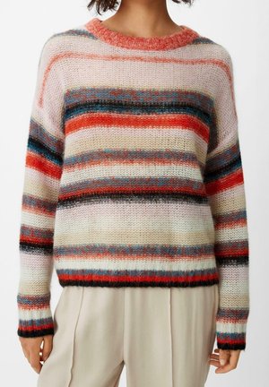 Woman wearing a fuzzy knit sweater with horizontal multicolored stripes in red, blue, black, white, and beige, paired with light beige pants.