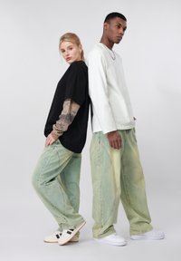 Ένας άνδρας και μια γυναίκα στέκονται πλάτη με πλάτη φορώντας φαρδιά ανοιχτοπράσινα τζιν; αυτός φοράει λευκό πουκάμισο με μακριά μανίκια, αυτή φοράει μαύρο φαρδύ μπλουζάκι.