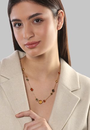 Halskette in Goldton mit runden Perlen in Bernstein, Braun und Olivgrün, präsentiert an einem Modell, das einen beigen Mantel trägt.