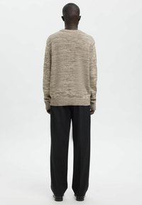 Beiger, strukturierter Pullover mit geripptem Saum und Bündchen, kombiniert mit schwarzen, weit geschnittenen Hosen, von hinten vor einem schlichten Hintergrund zu sehen.