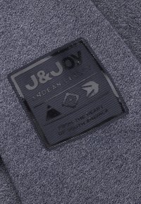 Donkergrijze stofpatch met reliëf logo-details, inclusief tekst en symbolen, die het merk en de onderhoudsinstructies schetsen, met een zachte textuur.