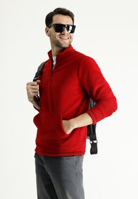 Suéter de forro polar rojo con cuello de cremallera y bolsillo frontal, combinado con pantalones grises y una mochila negra, accesorio con gafas de sol negras.