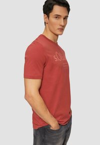 Rotes Baumwoll-T-Shirt mit Rundhalsausschnitt und kurzen Ärmeln, mit einem dezenten Logo-Print auf der Vorderseite. Kombiniert mit dunkelgrauen Jeans.
