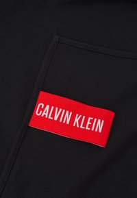 Černá tkanina s texturovaným povrchem, s výraznou červenou etiketou se bílým textem "CALVIN KLEIN" přišitým na kapse.