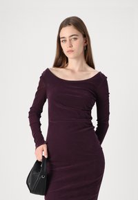 Deep purple long-sleeve dress with a wide neckline, gathered detail at the shoulder, and a fitted silhouette, paired with a small black handbag.